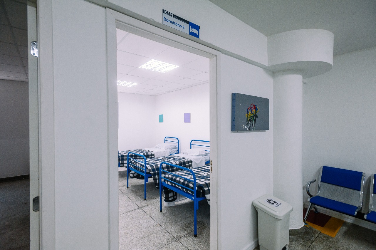 Na imagem mostra uma sala aberta com camas enfileiras. As camas são azuis e estão cobertas com um cobertor xadrez.